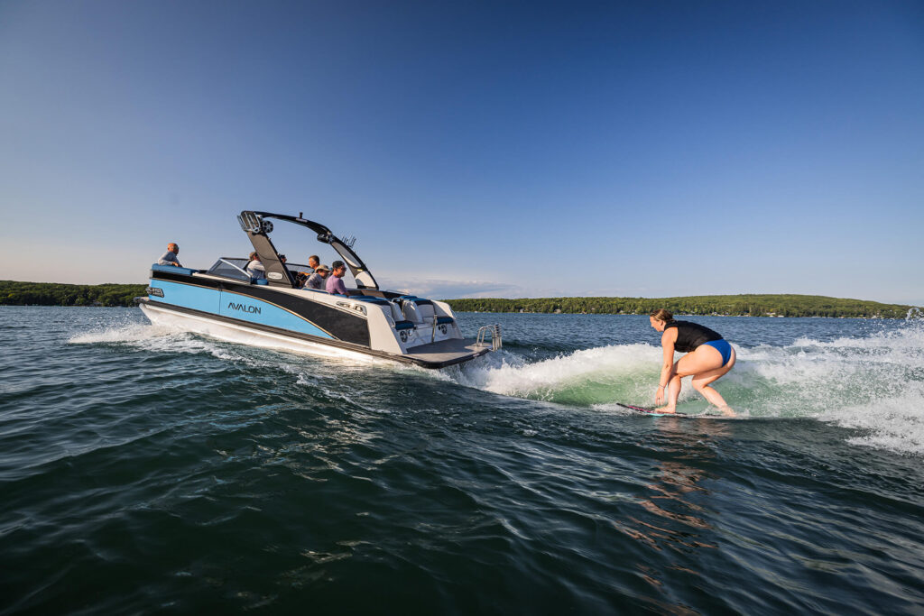 A person is wakesurfing behind a motorboat with several people onboard. The boat is sleek and modern, gliding over calm blue water under a clear sky. Green hills are visible in the distance, indicating a serene lakeside setting.