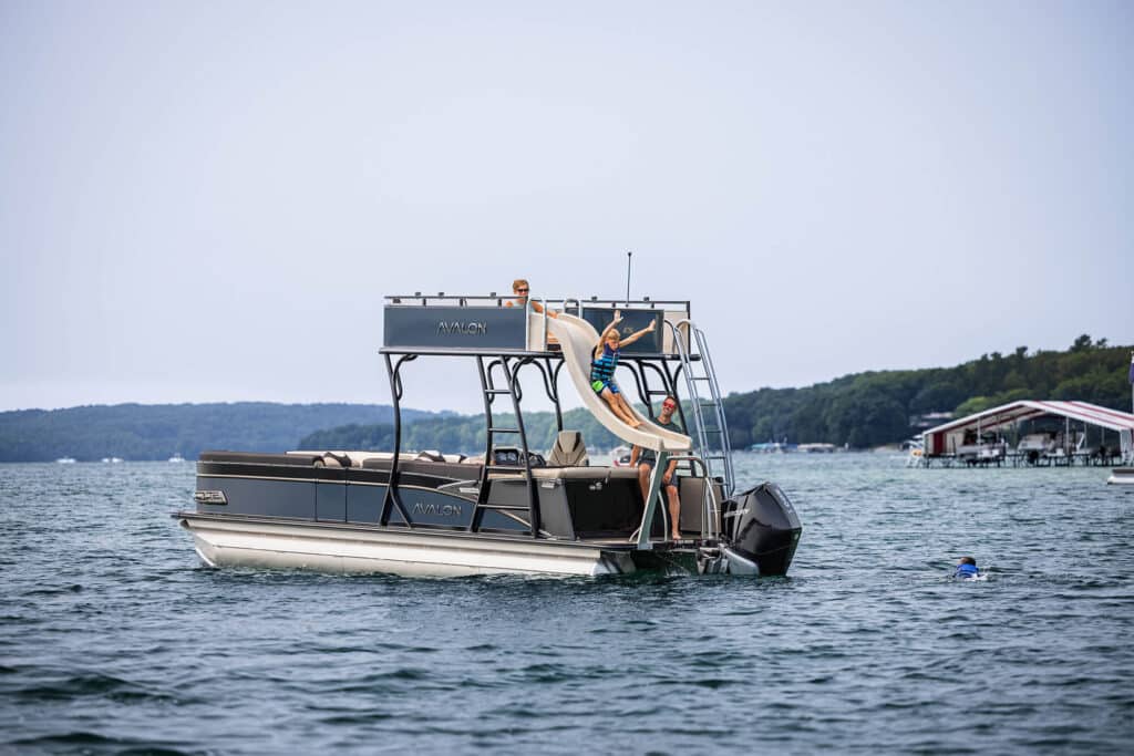 A pontoon boat equipped with a slide floats on a lake. A child is using the slide, while another person is in the water nearby. The boat is surrounded by calm waters with a shoreline and a distant dock visible in the background.