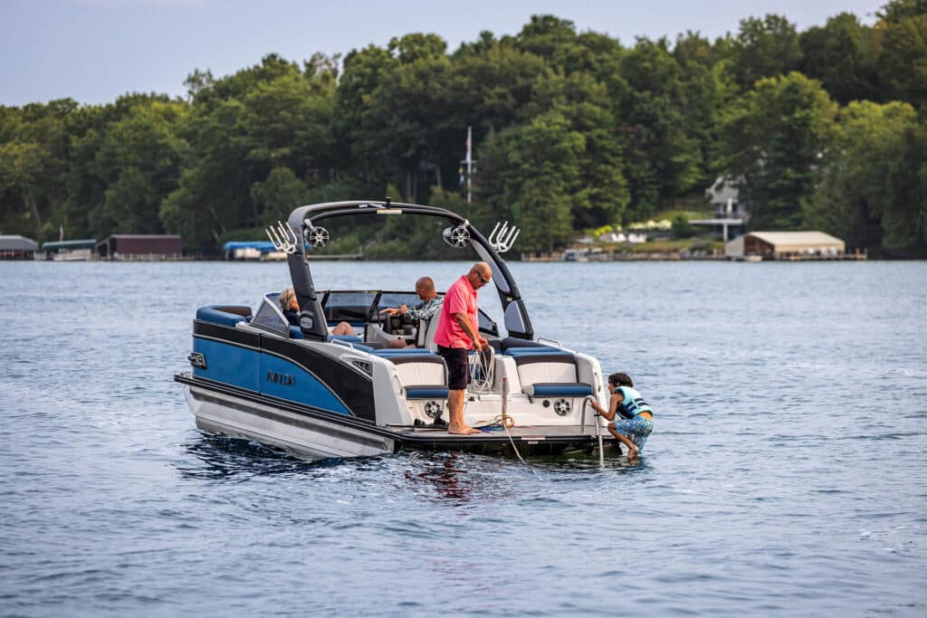 A blue and white motorboat is anchored in a calm lake. A man and a boy in life vests are on the boat's swim platform, with the boy climbing a ladder from the water. Surrounding them are lush green trees and several waterfront buildings in the background.