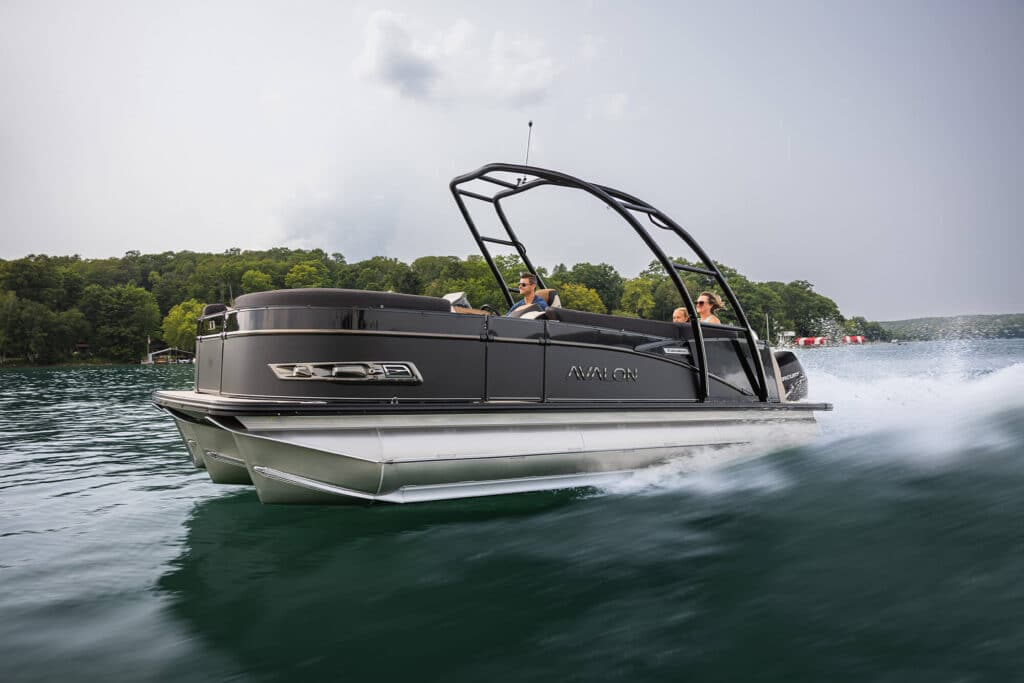 A sleek, black Avalon pontoon boat speeds across a calm lake. Three people are on board, enjoying the ride. The surrounding landscape features lush, green, tree-covered hills under a cloudy sky. The water splashes as the boat moves swiftly.
