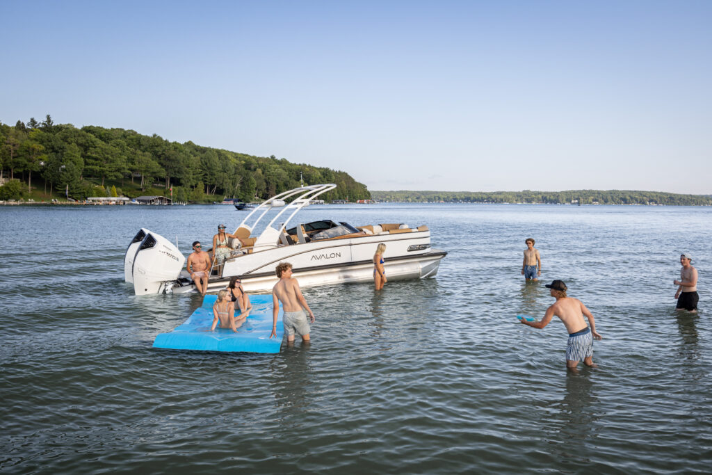 A group of people enjoying a sunny day on a lake. Some are swimming around a white Catalina Cruise Rear Bench Windshield pontoon boat, while others relax on a floating mat. The background features a wooded shoreline under a clear blue sky.